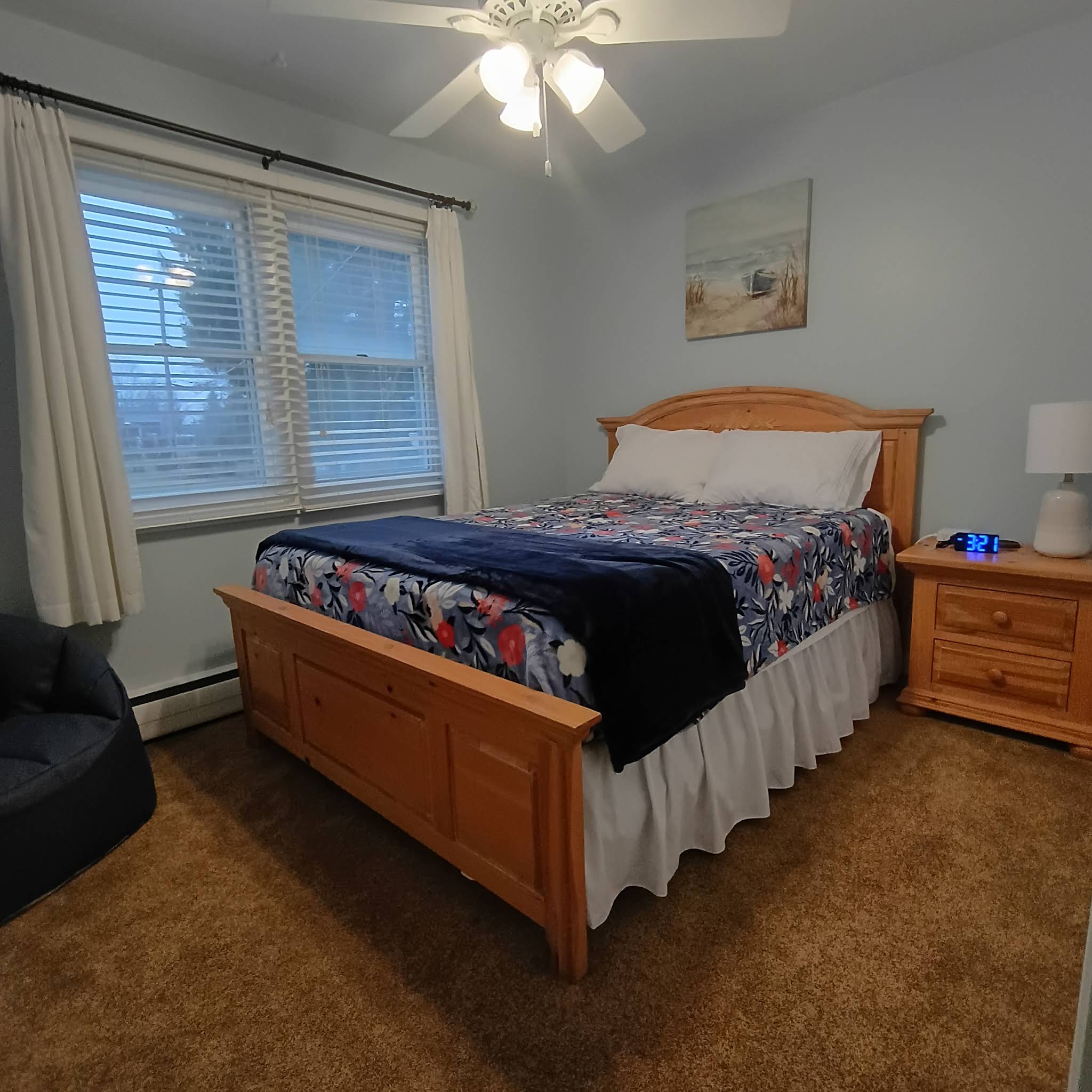 Bright bedroom with a queen bed, floral bedding, ceiling fan, and reading chair